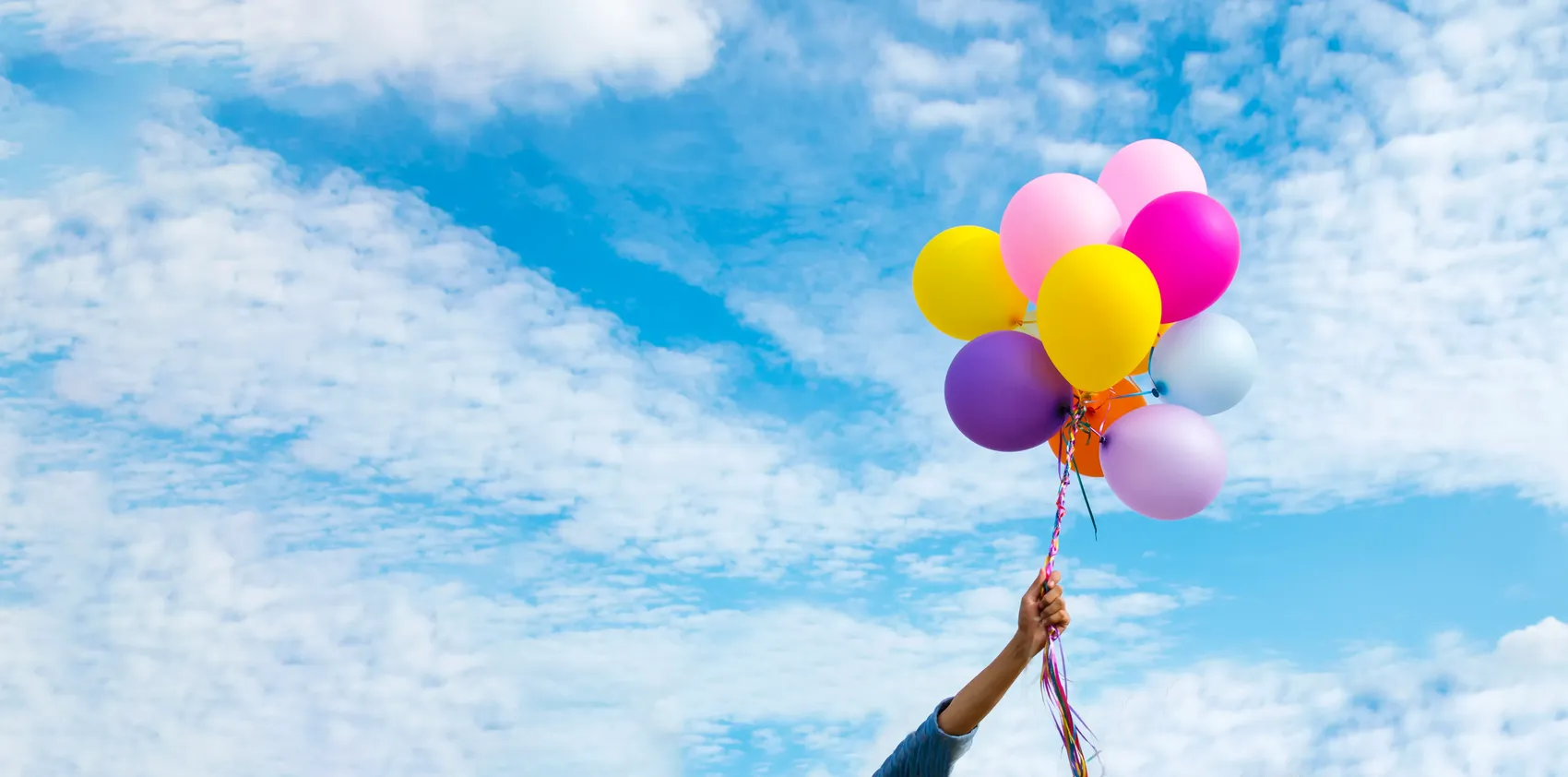 Bunte Luftballons steigen in einen blauen Himmel mit Wolken auf, gehalten von einer Hand.