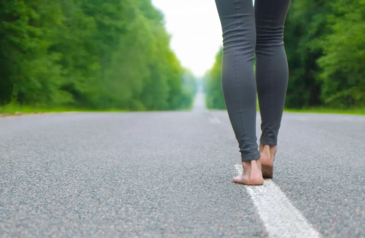 Barfüßige Person geht auf einer weißen Linie auf einer asphaltierten Straße durch eine grüne Waldlandschaft.