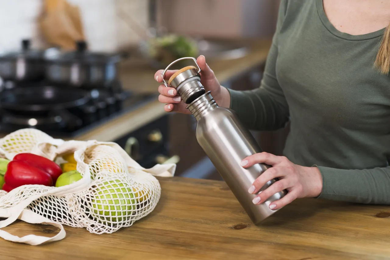 Eine Person hält eine silberne Edelstahl-Trinkflasche in einer Küche neben einer Netztasche mit Obst und Gemüse.
