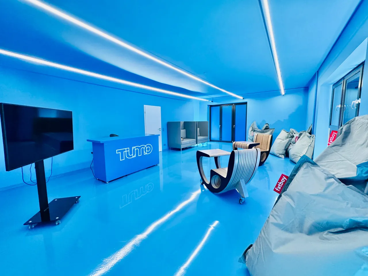 Heller, moderner Raum mit blauen Wänden und reflektierendem Boden. Möbelstücke wie Beanbags, Sessel und ein Schreibtisch mit Logo sind zu sehen.
