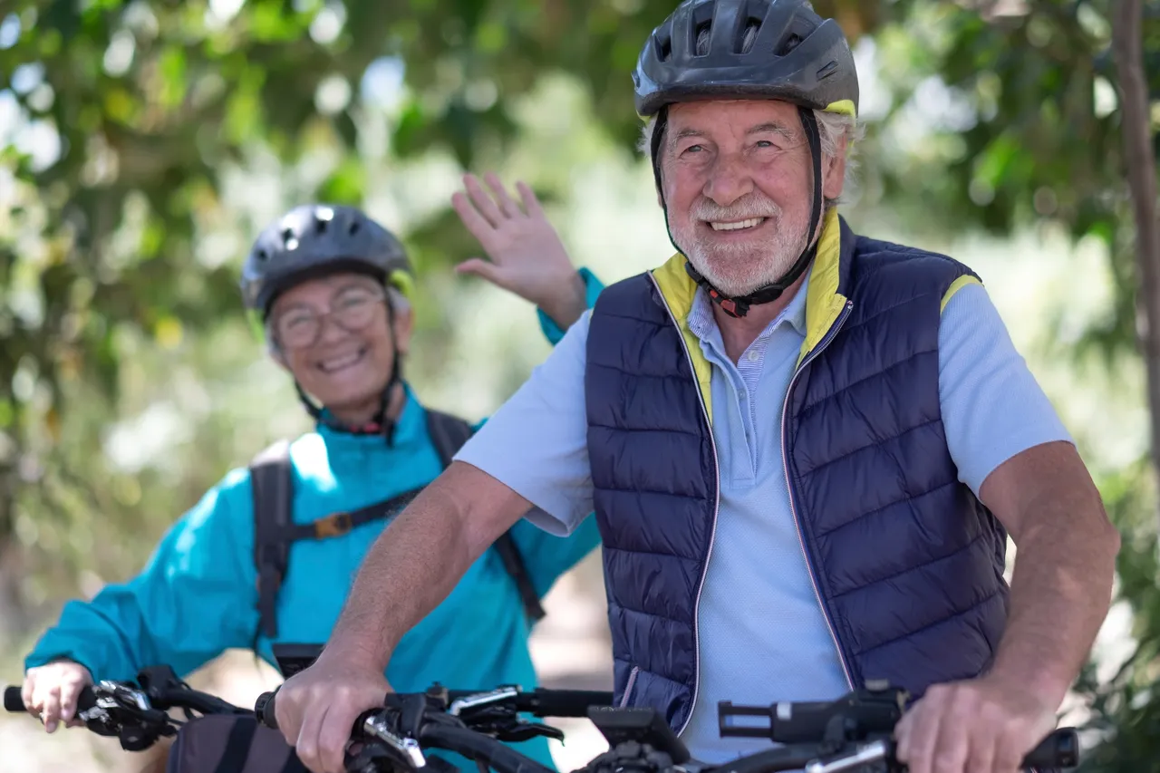 Ein älterer Mann mit Helm winkt lächelnd in die Kamera, während er auf einem E-Bike fährt. Im Hintergrund ist eine ältere Frau auf einem weiteren E-Bike zu sehen.