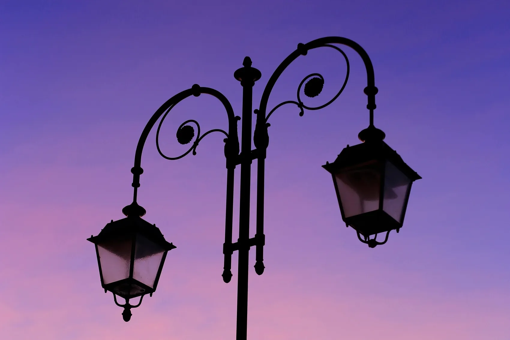 Silhouette einer verzierten, zweiflammigen Laterne vor einem violett-rosafarbenen Himmel.