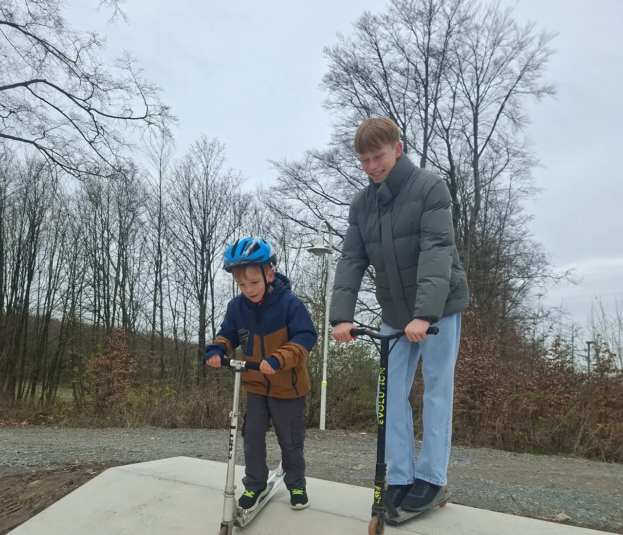 Zwei Jungen, ein älterer und ein jüngerer, stehen auf einer Betonrampe und fahren mit ihren Scootern. Im Hintergrund sind kahle Bäume zu sehen.