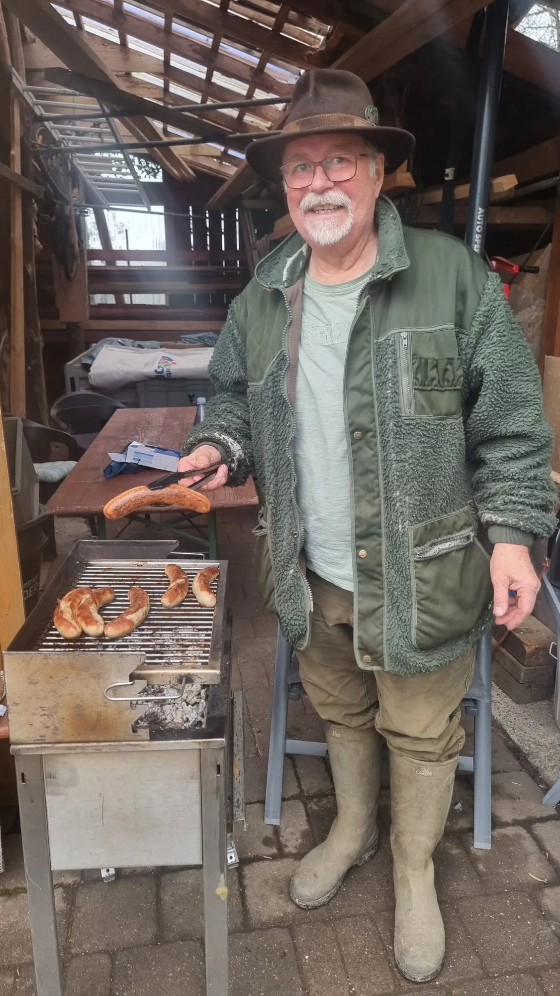 Ein Mann mit Hut und Jacke steht lächelnd vor einem Grill mit Bratwürsten und Brötchen. Im Hintergrund sind Tische und Stühle zu sehen.