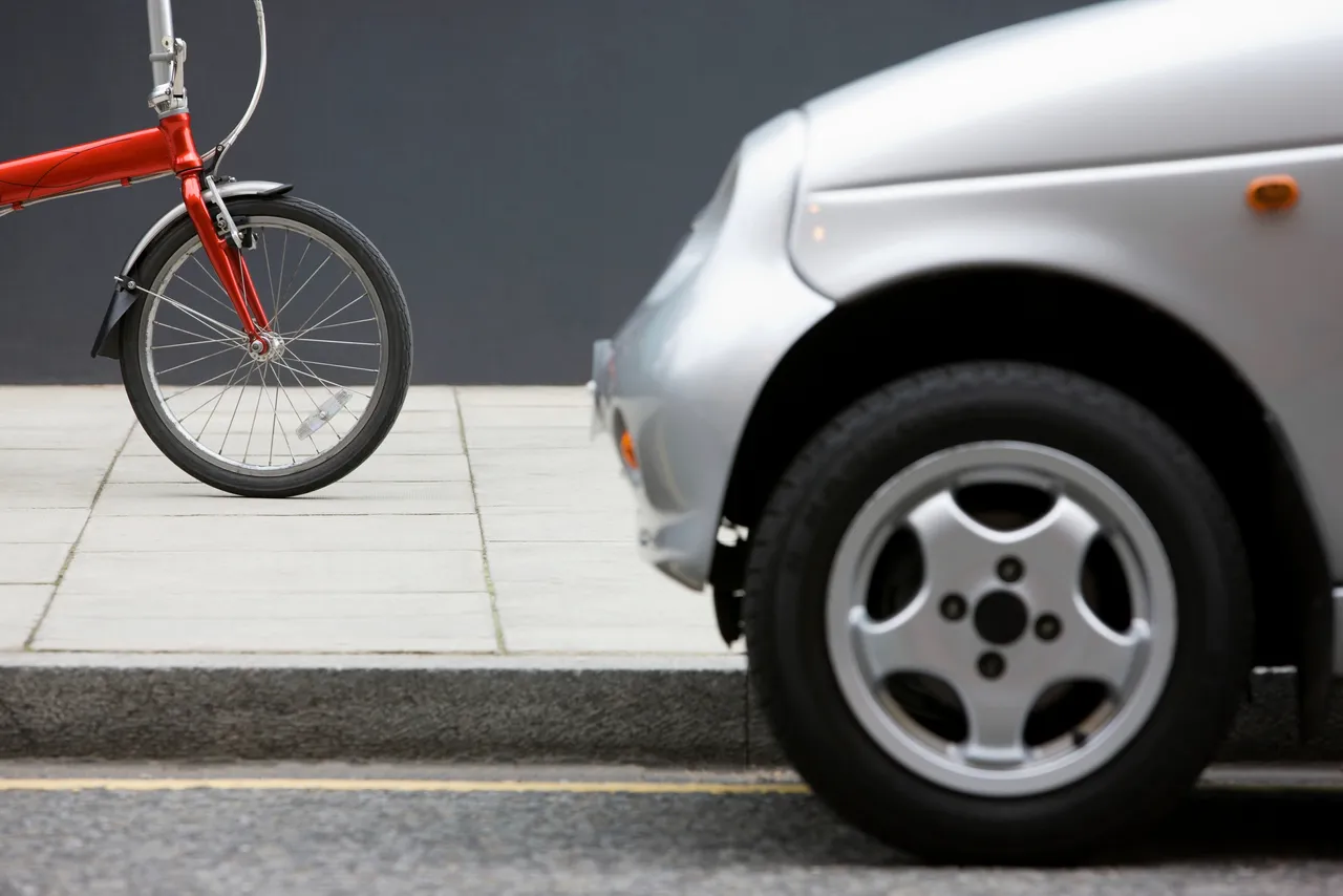 Nahaufnahme eines roten Fahrradrades auf dem Gehweg neben einem silbernen Autoreifen am Straßenrand.