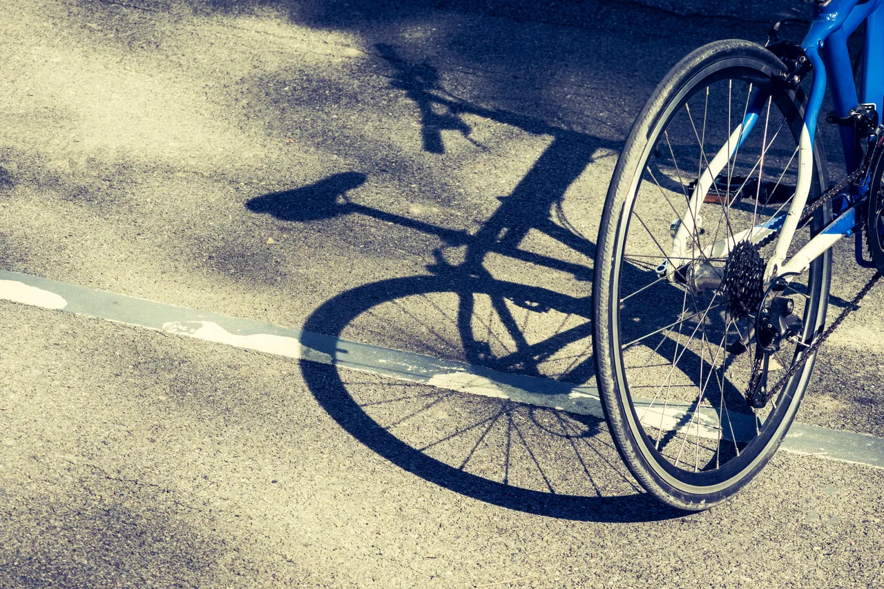 Nahaufnahme eines blauen Fahrradrads und seines Schattens auf einem asphaltierten Radweg.