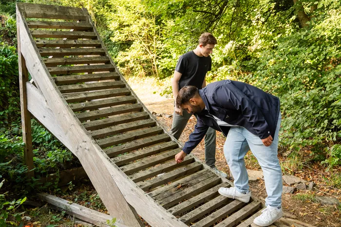 Zwei junge Männer begutachten eine hölzerne Rampe in einem Waldgebiet. Einer der Männer berührt dabei die Holzlatten der Konstruktion.