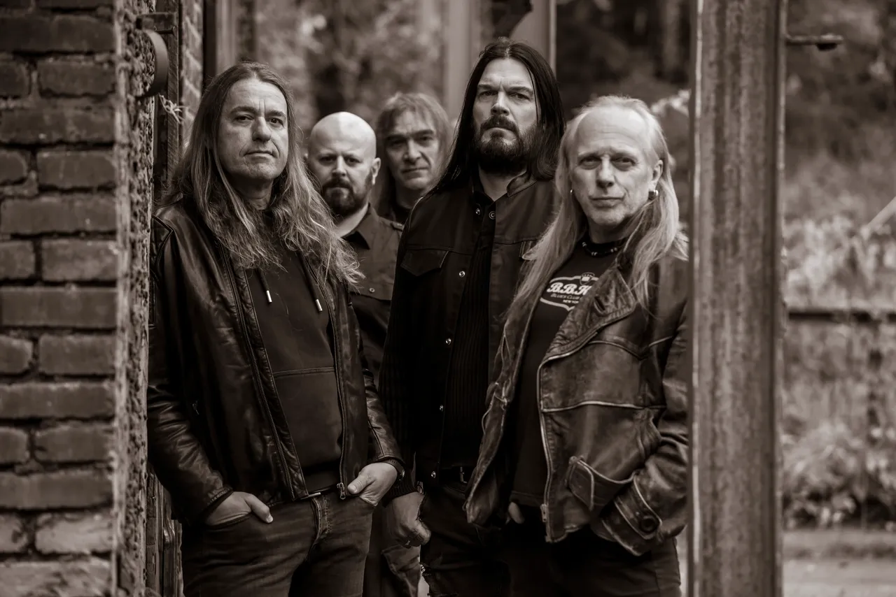 Fünf Männer, die Band „Bonded“, posieren vor einer verfallenen Backsteinmauer und einem Holztor. Sie tragen dunkle Kleidung und haben lange Haare.