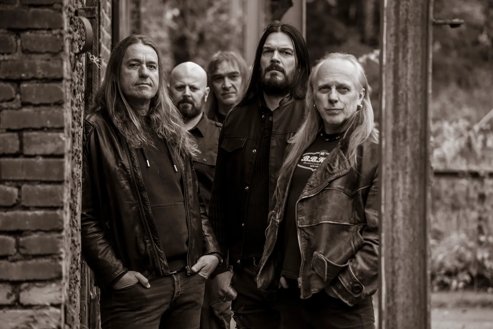 Fünf Männer, die Band „Bonded“, posieren vor einer verfallenen Backsteinmauer und einem Holztor. Sie tragen dunkle Kleidung und haben lange Haare.