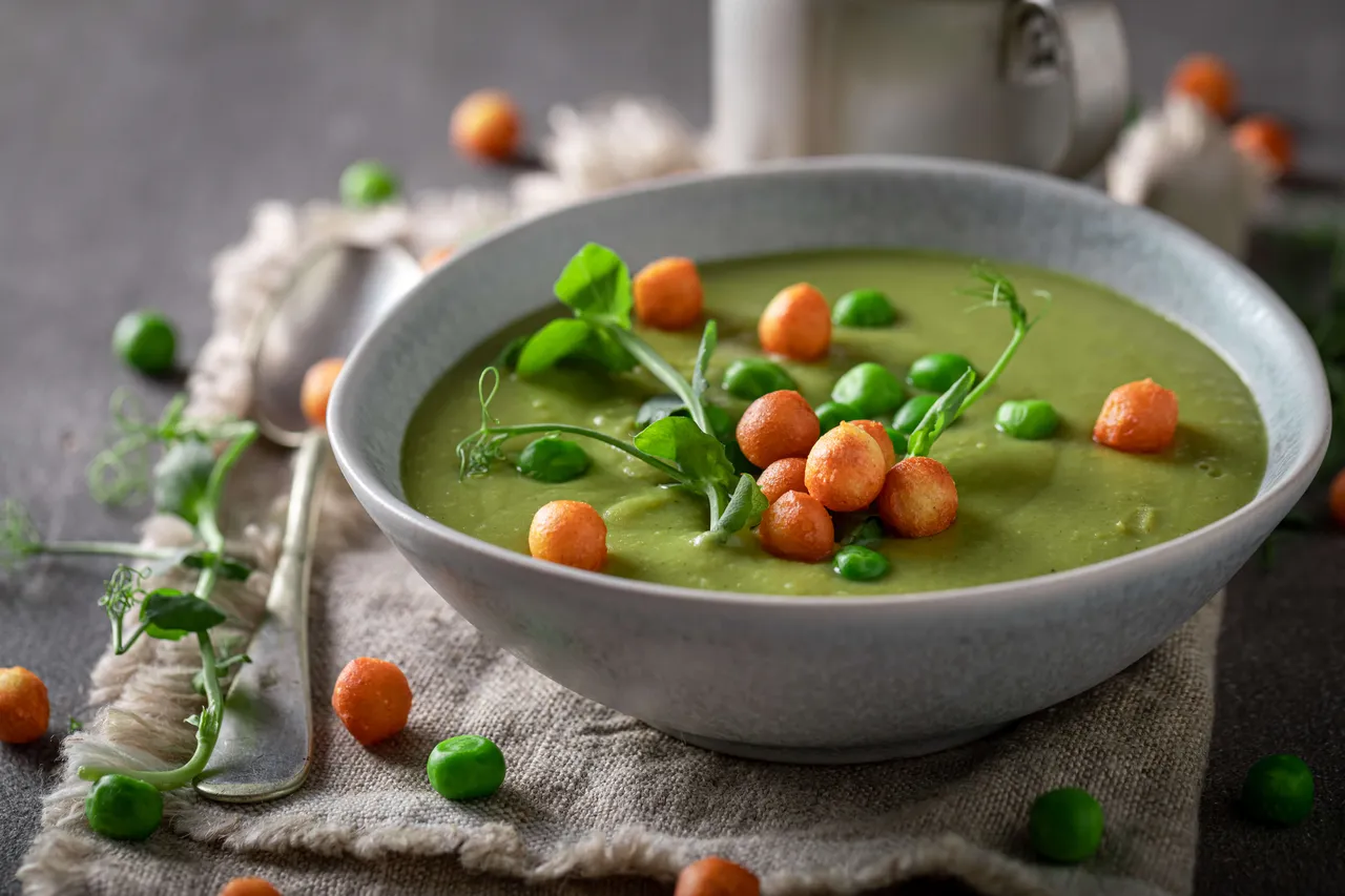 Grüne Erbsensuppe in einer hellgrauen Schüssel, garniert mit orangefarbenen Croutons, ganzen Erbsen und frischen Erbsensprossen.