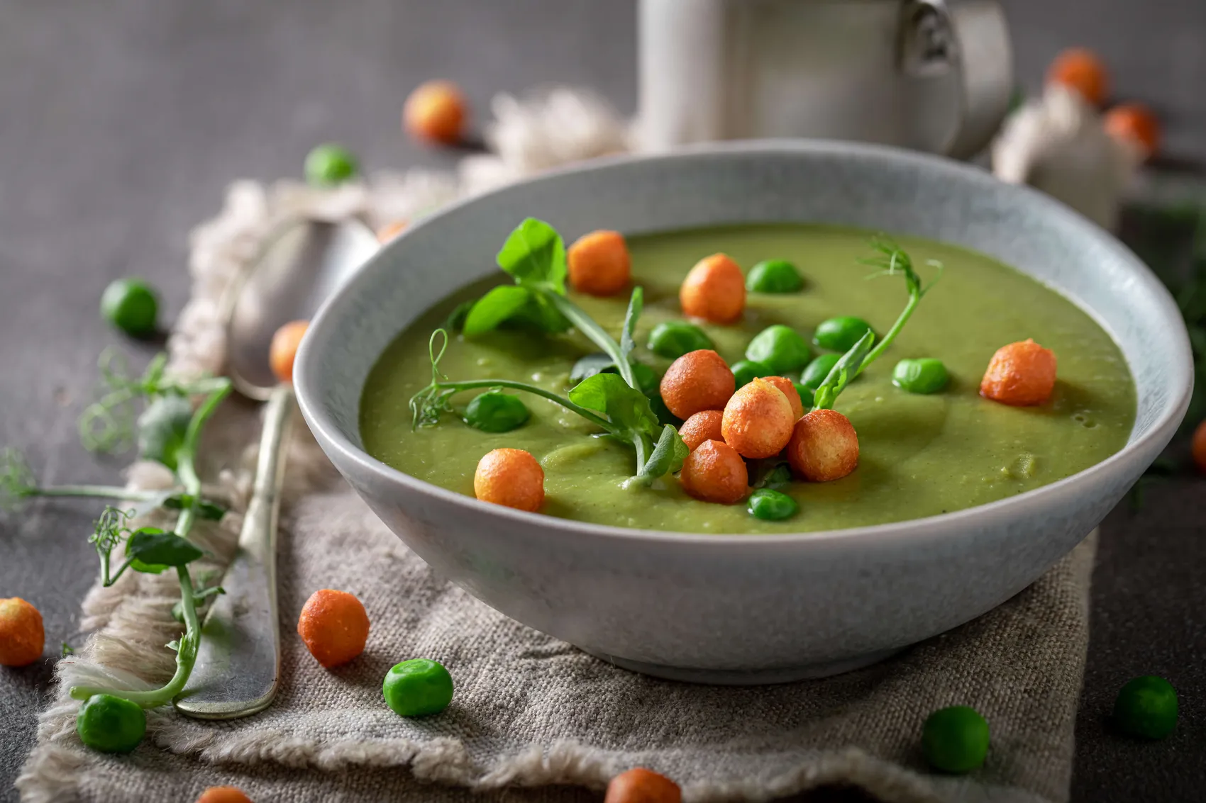 Grüne Erbsensuppe in einer hellgrauen Schüssel, garniert mit orangefarbenen Croutons, ganzen Erbsen und frischen Erbsensprossen.