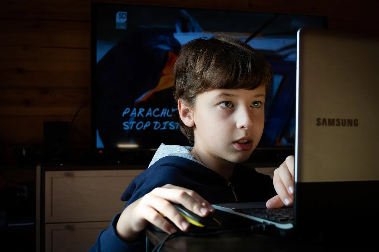 Junge konzentriert sich beim Bedienen eines Computers, im Hintergrund ein Fernseher mit einer Szene von Fallschirmspringern und dem Text „Parachute Stop Dis“. Das Bild illustriert das Thema Medienprävention für Kinder.