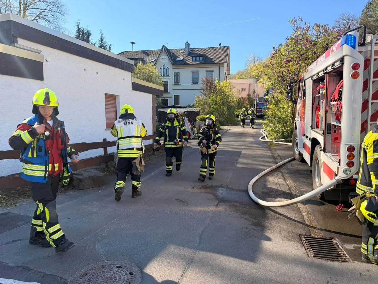 Feuerwehrleute in voller Montur gehen auf der Straße an einem roten Feuerwehrauto vorbei, während im Hintergrund ein Gebäude steht.