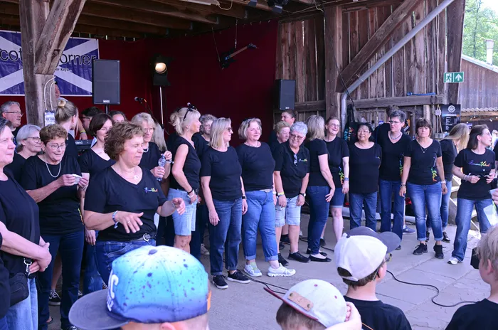 Große Gruppe von Frauen in schwarzen T-Shirts und Jeans steht auf einer Bühne und singt oder tanzt beim I-Rock Festival in Plettenberg.