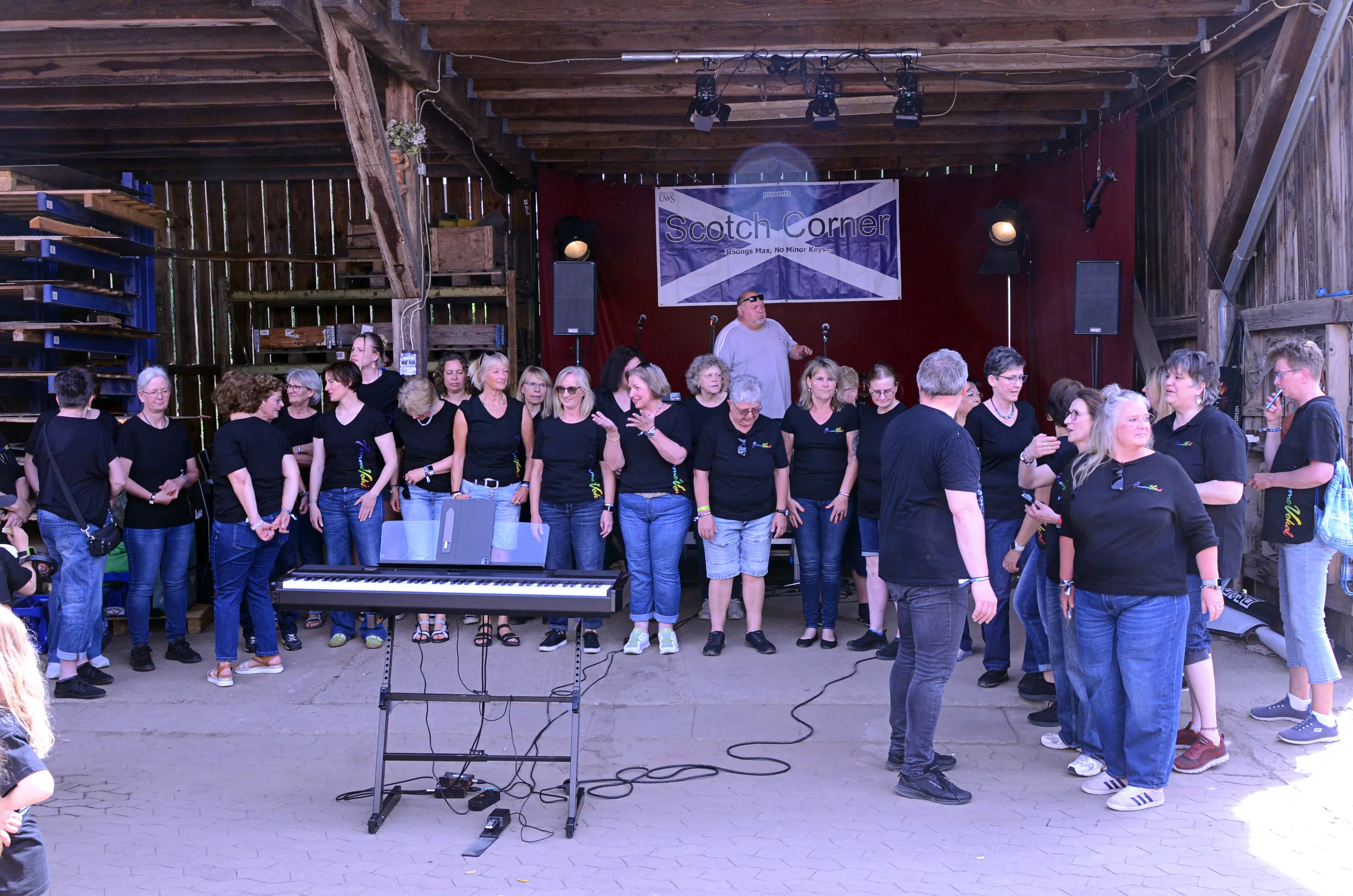 Chor „Femme Vocal“ singt auf der Bühne des I-Rock Festivals immecke in Plettenberg. Die Sängerinnen und Sänger stehen vor einem Banner mit der Aufschrift „Scotch Corner“ und werden von einem Mann mit Keyboard begleitet.