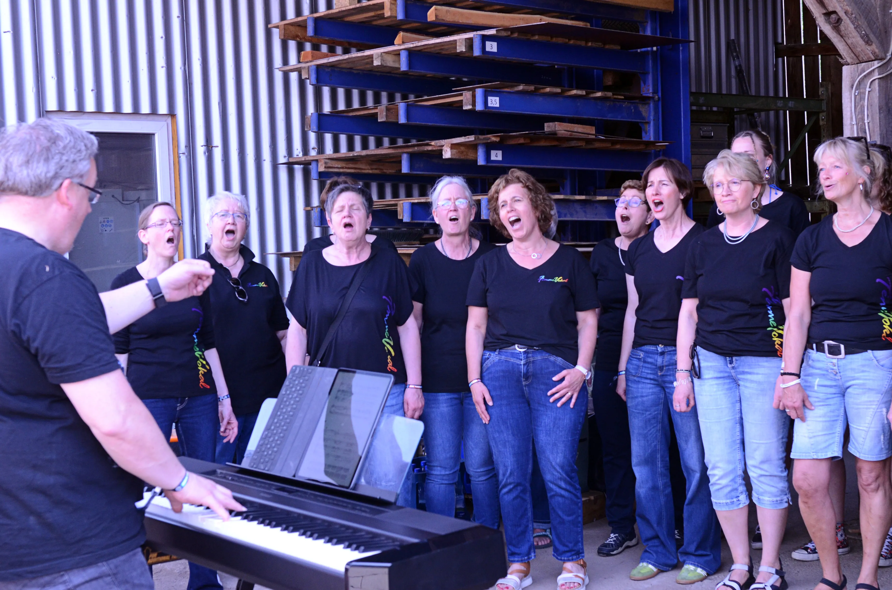 Chor singt vor einem Lagerhaus mit Palettenregalen. Ein Mann spielt Klavier und dirigiert den Chor, dessen Mitglieder hauptsächlich Frauen mittleren Alters sind.