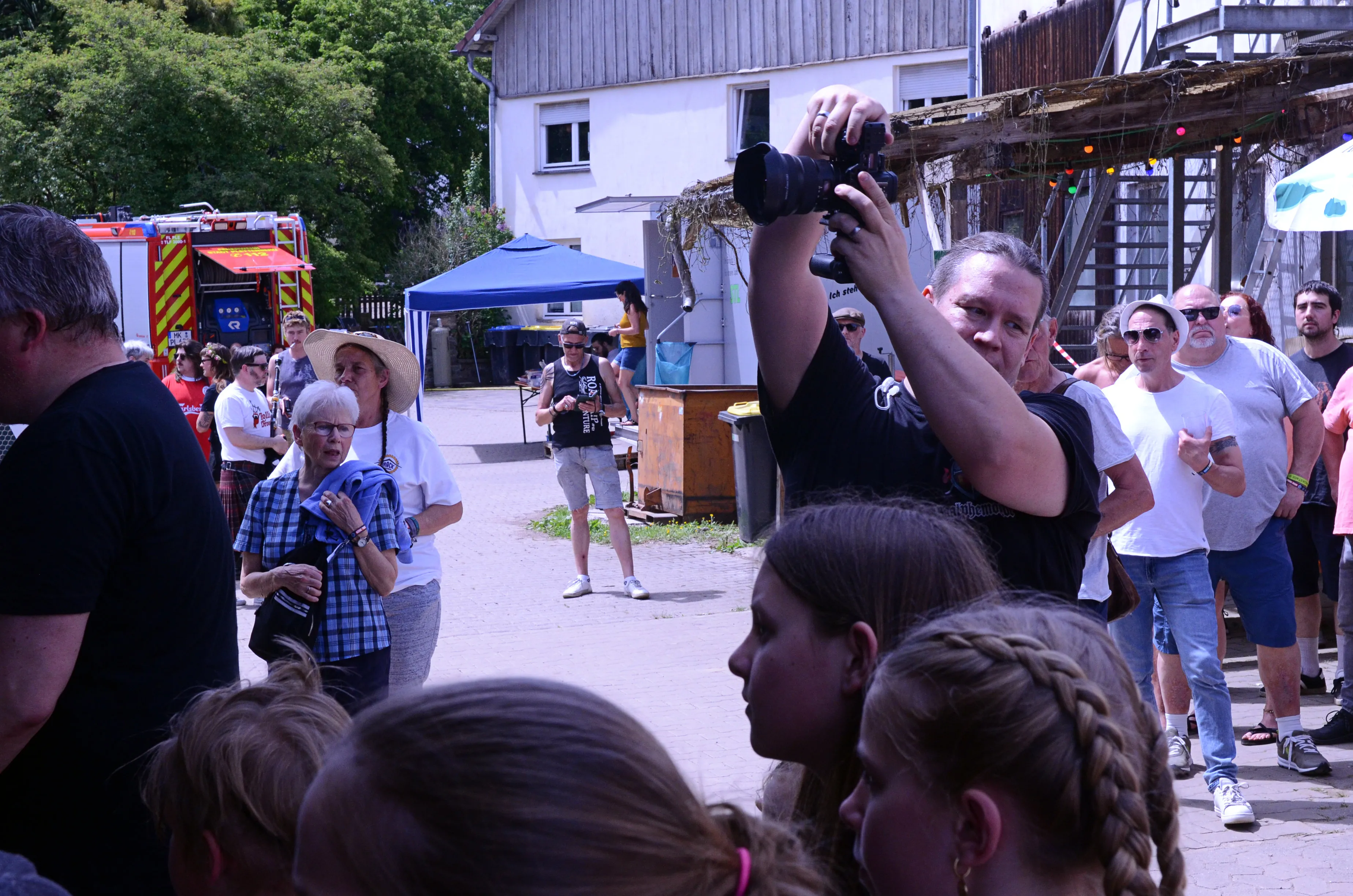 Ein Mann fotografiert mit einer professionellen Kamera eine Menschenmenge bei einem Open-Air-Festival. Im Hintergrund sind weitere Festivalbesucher und ein rotes Feuerwehrfahrzeug zu sehen.