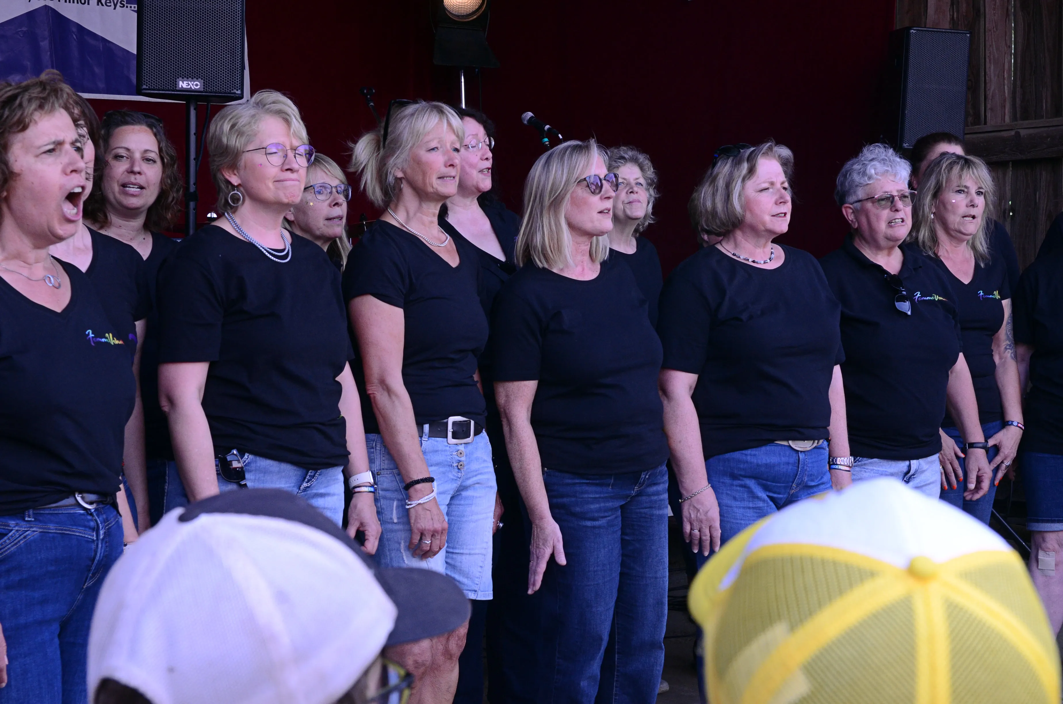 Chor aus mehreren Frauen singt auf einer Bühne, die mit Lautsprechern ausgestattet ist. Die Sängerinnen tragen schwarze T-Shirts und Jeans.