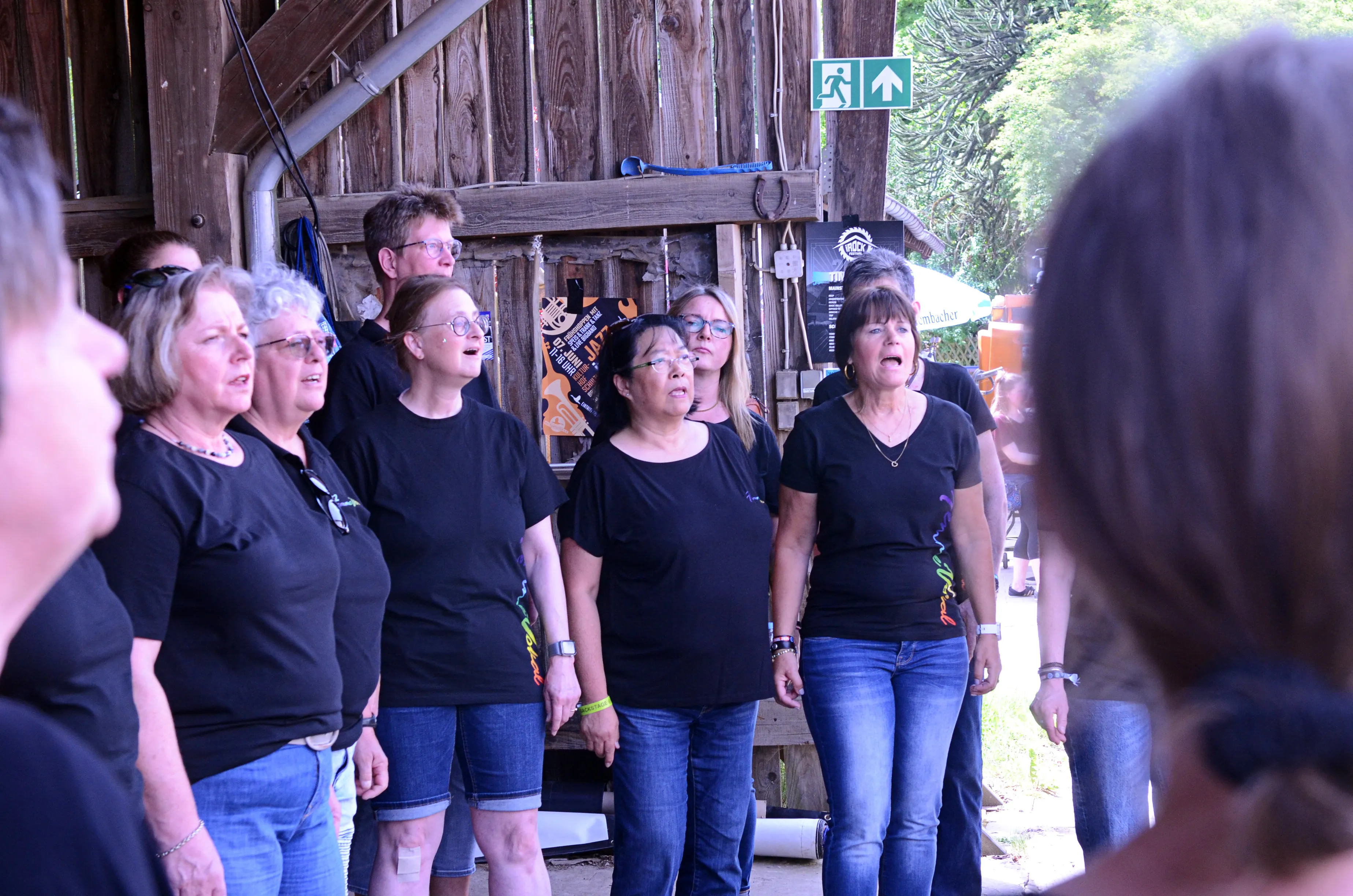 Chor singt auf der Bühne des I-Rock Festivals in Plettenberg. Die Gruppe besteht aus mehreren Frauen und Männern, die schwarze T-Shirts und Jeans tragen.