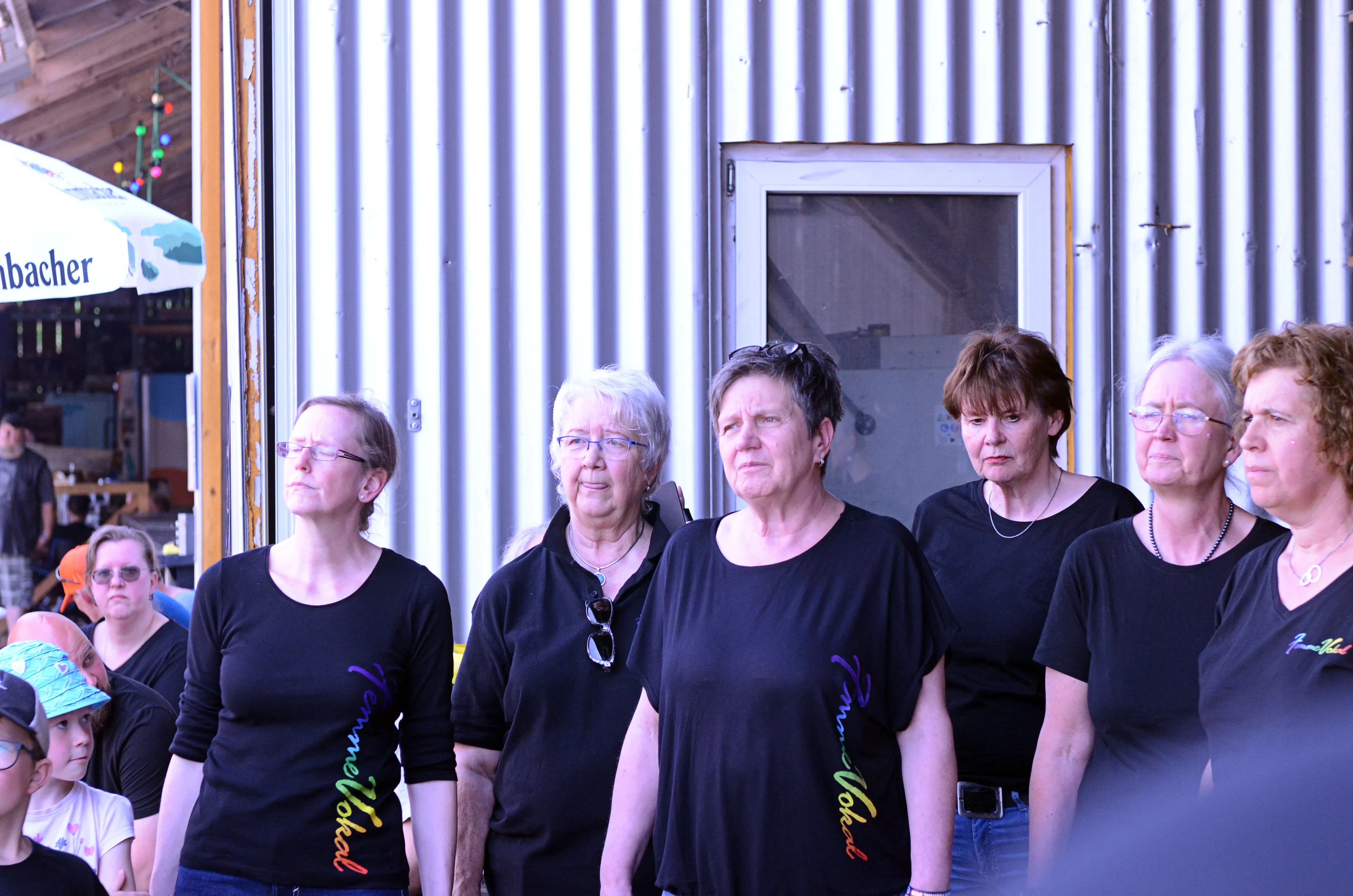 Fünf Frauen stehen eng nebeneinander und blicken nach oben, während sie schwarze T-Shirts mit Aufschrift tragen. Im Hintergrund ist ein Festivalgelände mit Zuschauern und einem Getränkestand zu sehen.