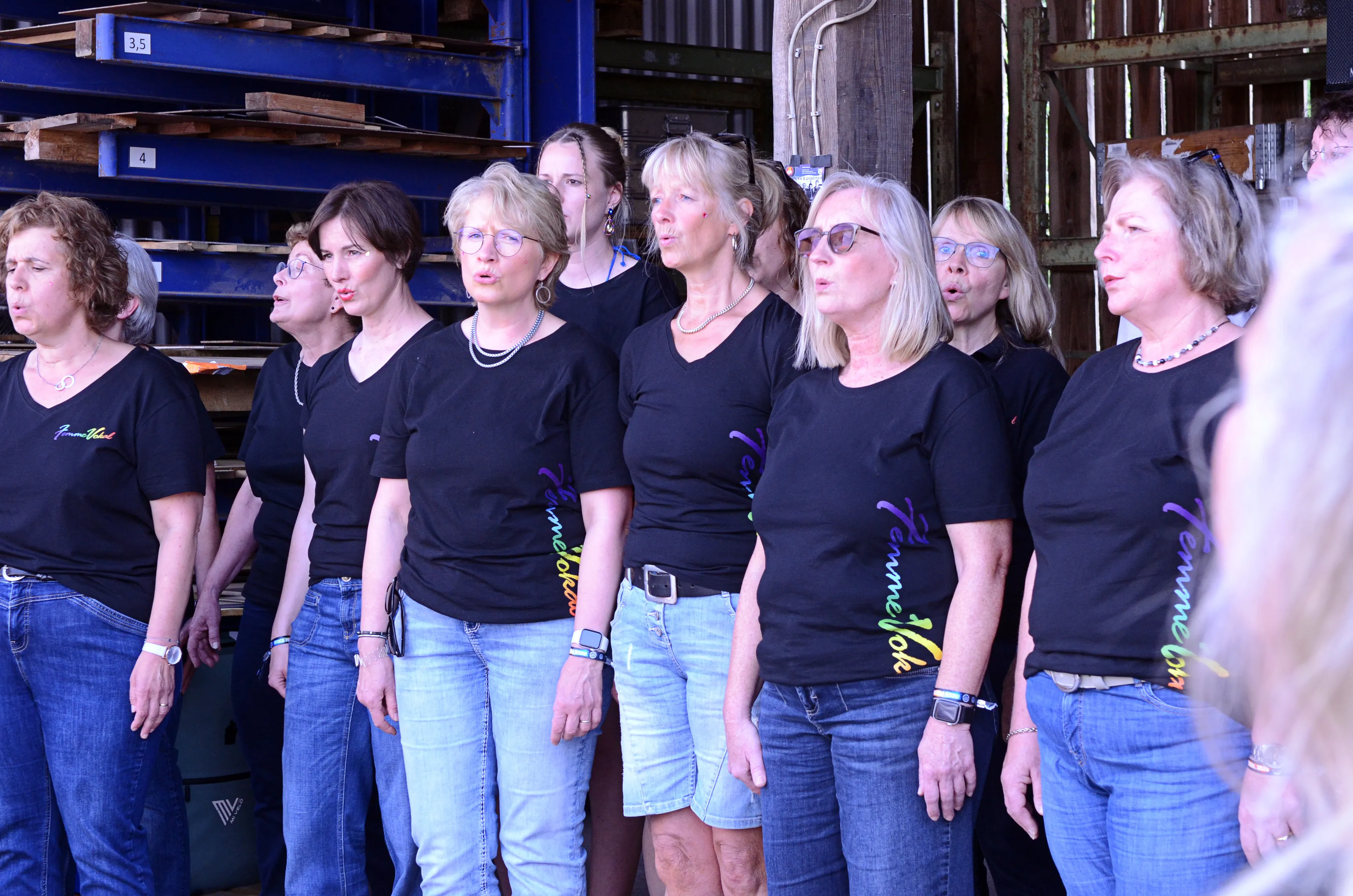 Chor „Femmevokal“ singt auf der Bühne des I-Rock Festivals in Plettenberg. Die Sängerinnen tragen schwarze T-Shirts mit violettem Aufdruck und Jeans.