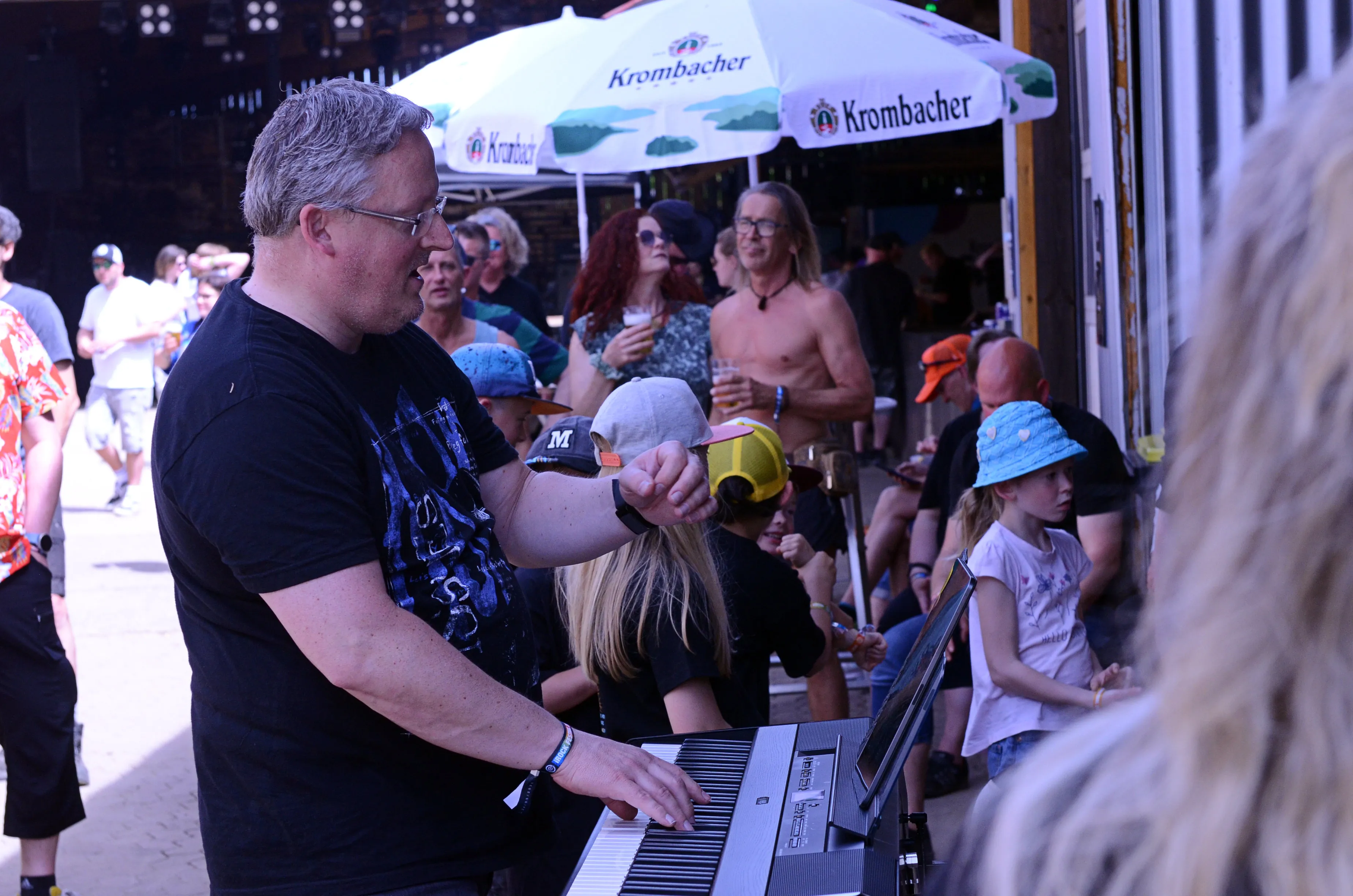 Ein Mann spielt Keyboard vor einer Menschenmenge auf dem I-Rock Festival in Plettenberg. Im Hintergrund sind Sonnenschirme mit dem Schriftzug „Krombacher“ zu sehen.