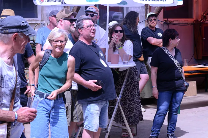 Zuschauer stehen vor einer Bühne im Freien, einige halten Getränke in der Hand. Im Hintergrund sind weitere Menschen und Sonnenschirme zu sehen.