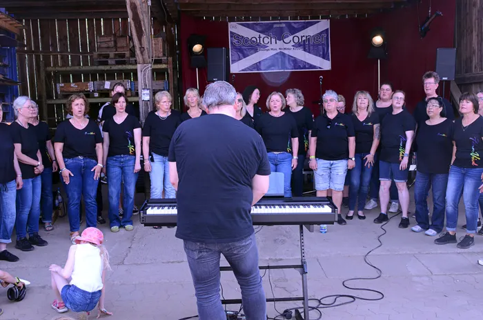 Chor mit rund 20 Personen, größtenteils Frauen, singt auf einer Bühne vor einem Banner mit dem Schriftzug „Scotch Corner“. Ein Mann dirigiert den Chor, während ein Kind im Vordergrund sitzt und zusieht.