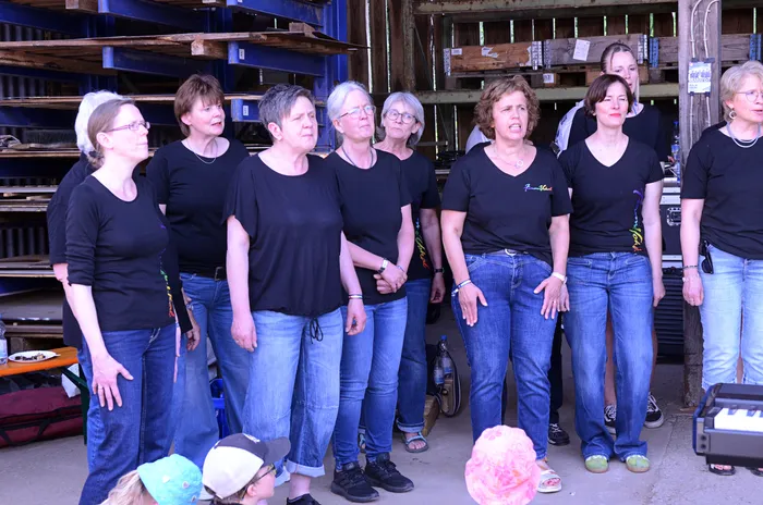Chorformation aus etwa zehn Frauen singt auf einer Bühne vor Holzwänden und Publikum. Sie tragen schwarze T-Shirts und Jeans.