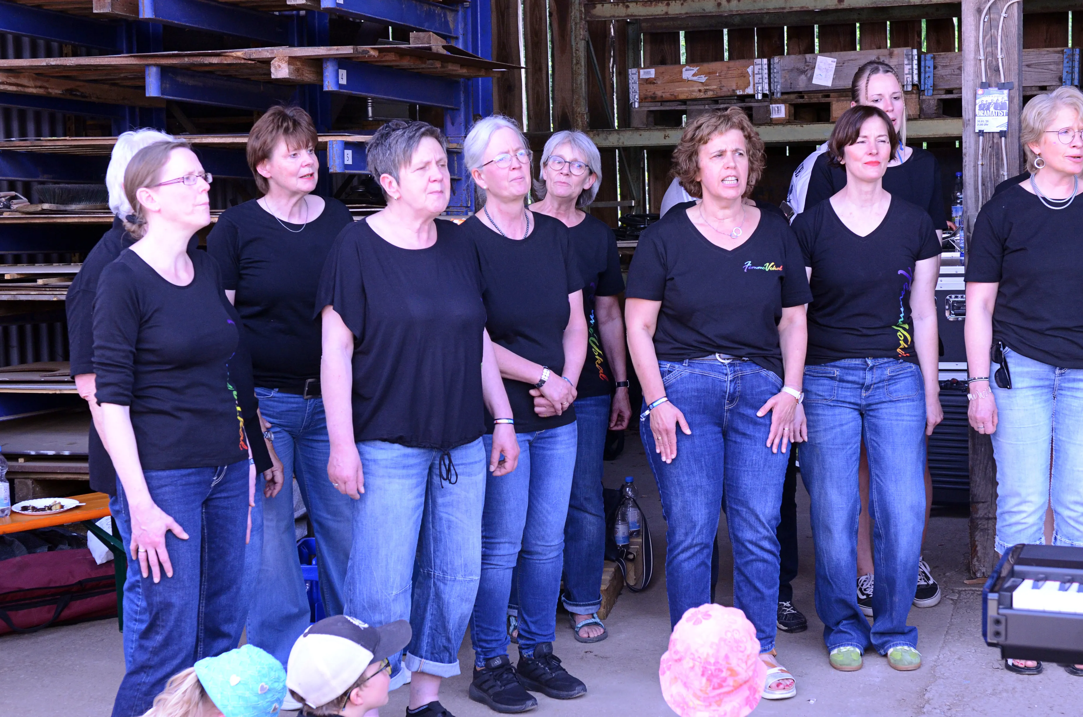 Chorformation aus etwa zehn Frauen singt auf einer Bühne vor Holzwänden und Publikum. Sie tragen schwarze T-Shirts und Jeans.