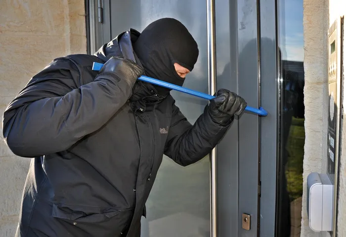 Eine Person mit dunkler Kleidung, Sturmhaube und Handschuhen versucht, mit einem blauen Hebelwerkzeug eine Glastür aufzuhebeln.