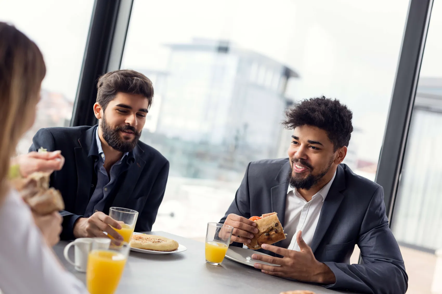 Drei Berufstätige in formeller Kleidung sitzen an einem Tisch und essen Sandwiches und trinken Orangensaft.