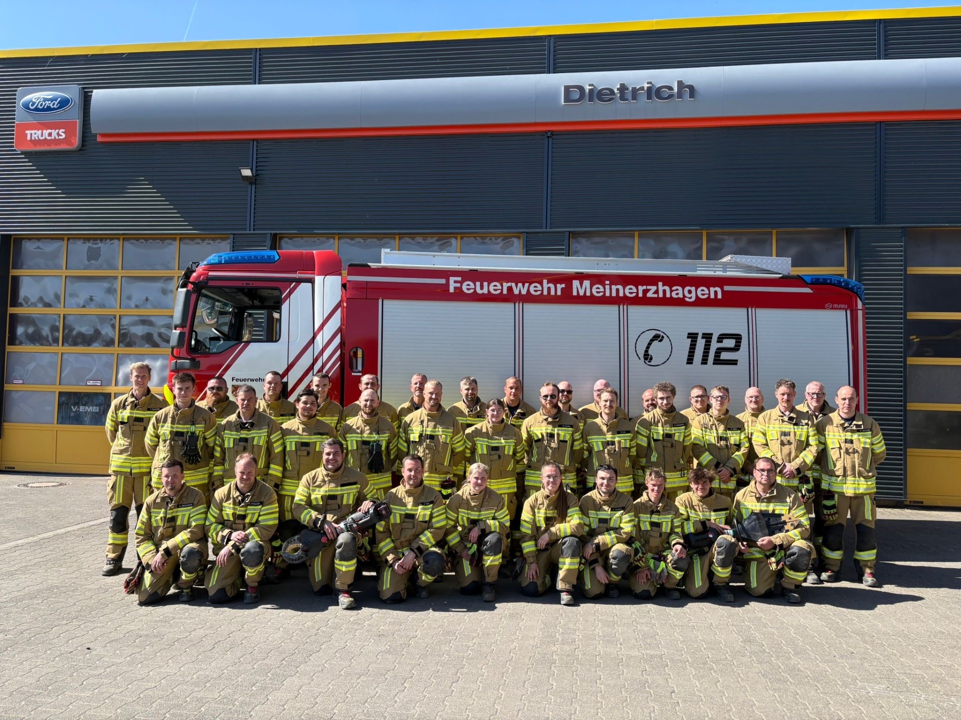 Gruppe von Feuerwehrleuten in voller Montur posiert vor einem roten Feuerwehrauto und dem Gebäude der Feuerwehr Meinerzhagen.