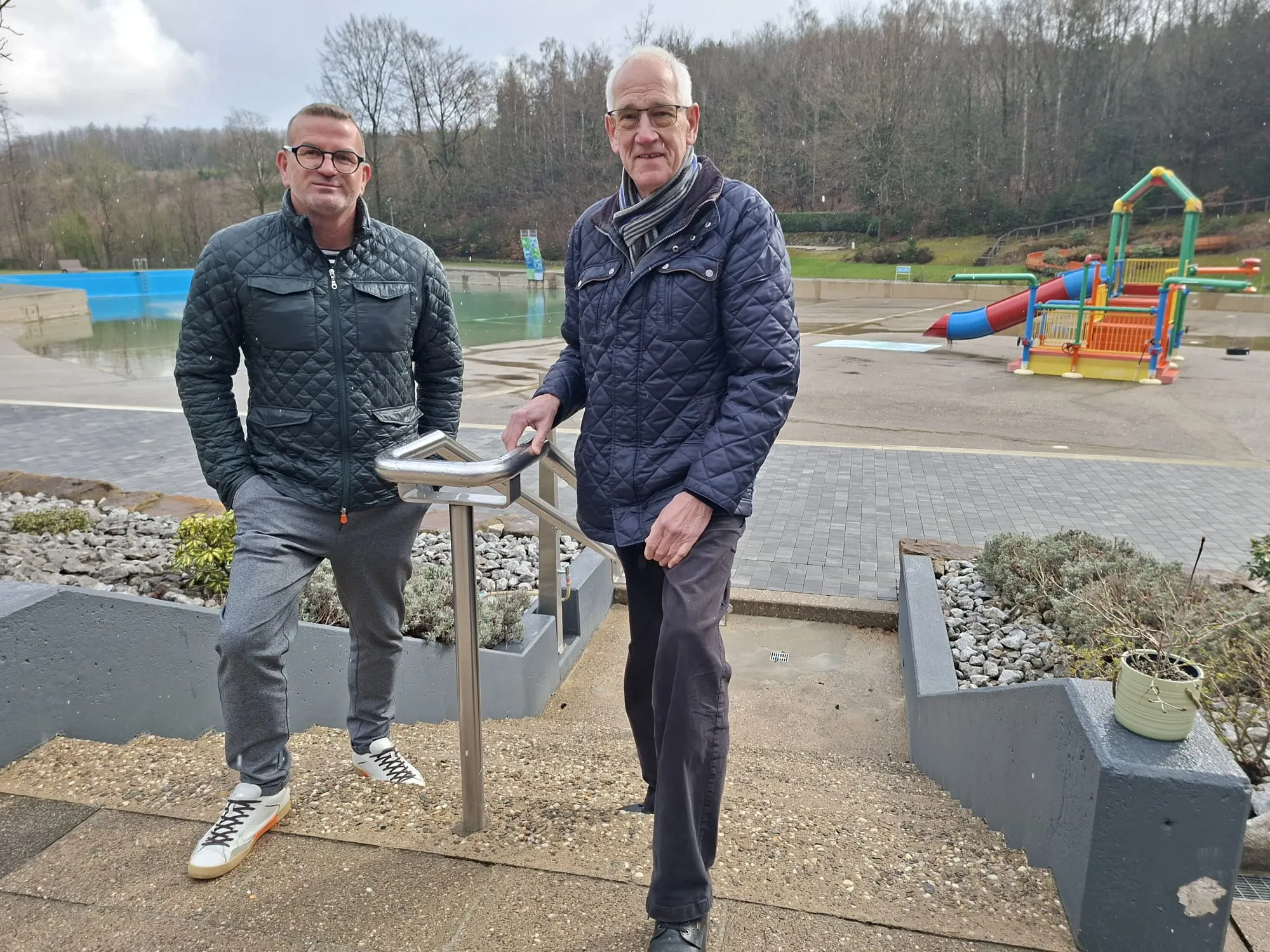 Zwei Männer stehen vor einem Becken mit einem Kinderspielplatz im Hintergrund. Sie scheinen die Bauarbeiten zu überwachen.
