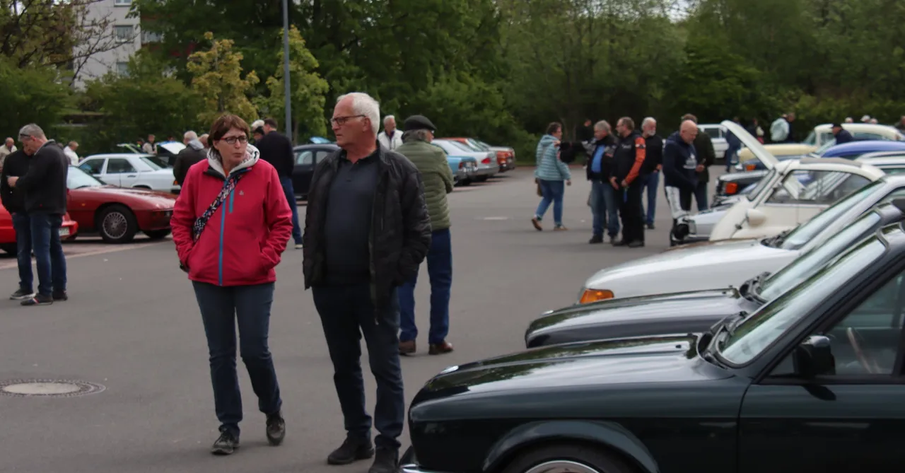 Zwei ältere Menschen gehen auf einem Parkplatz an einer Reihe von Oldtimern vorbei, während im Hintergrund weitere Besucher das Treffen betrachten.