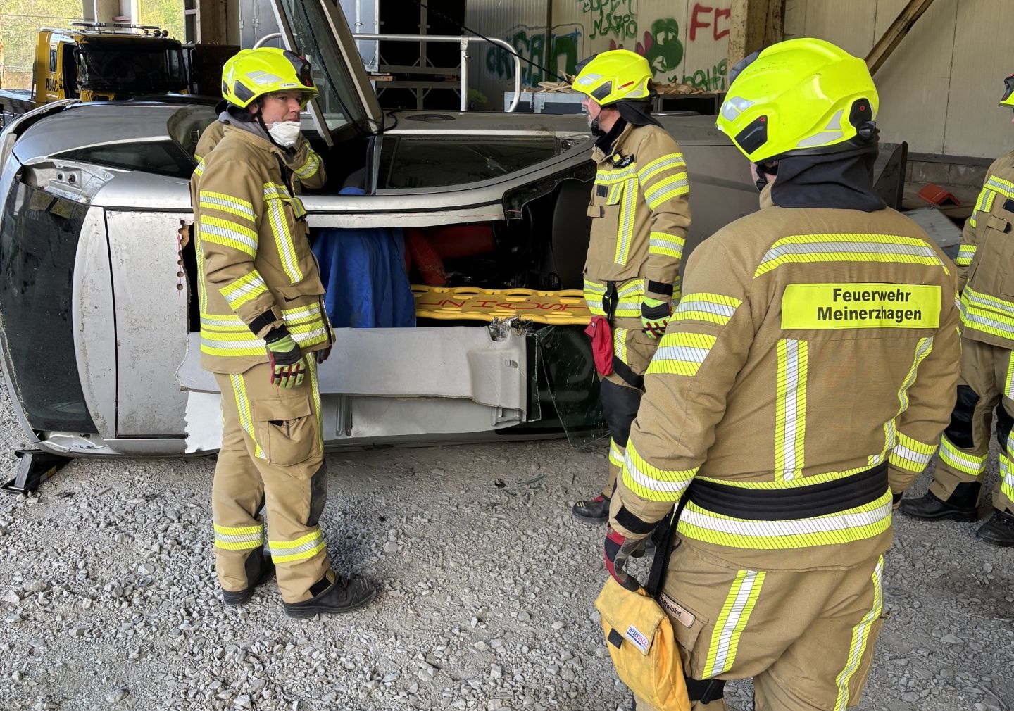 Vier Feuerwehrleute in voller Montur stehen um ein stark beschädigtes Auto, während sie sich beraten. Im Hintergrund ist ein LKW mit Graffiti zu sehen und der Schriftzug „Feuerwehr Meinerzhagen“.