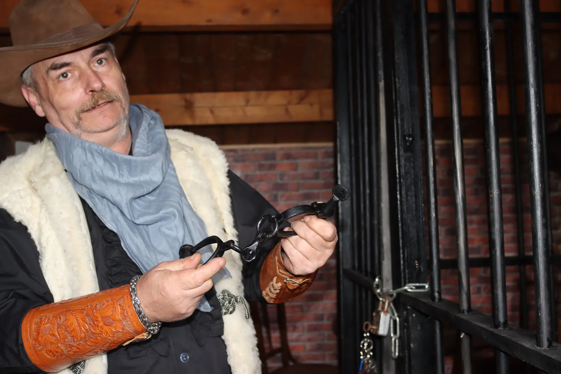 Ein Mann mit Cowboyhut und Halstuch hält Handschellen vor einem Gefängnisgitter. Im Hintergrund ist eine Backsteinmauer zu sehen.