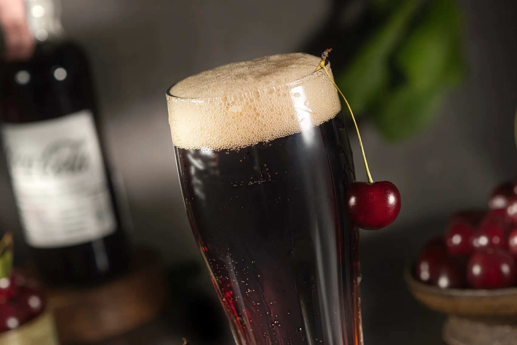 Ein Glas dunkles, kohlensäurehaltiges Getränk mit Kirsche am Rand und cremigem Schaum, vor dunklem Hintergrund und weiteren Kirschen.