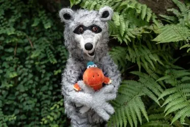 Ein grauer Waschbär aus Filz hält einen orangefarbenen Dino-Plüschtier mit blauer Mütze im Arm.