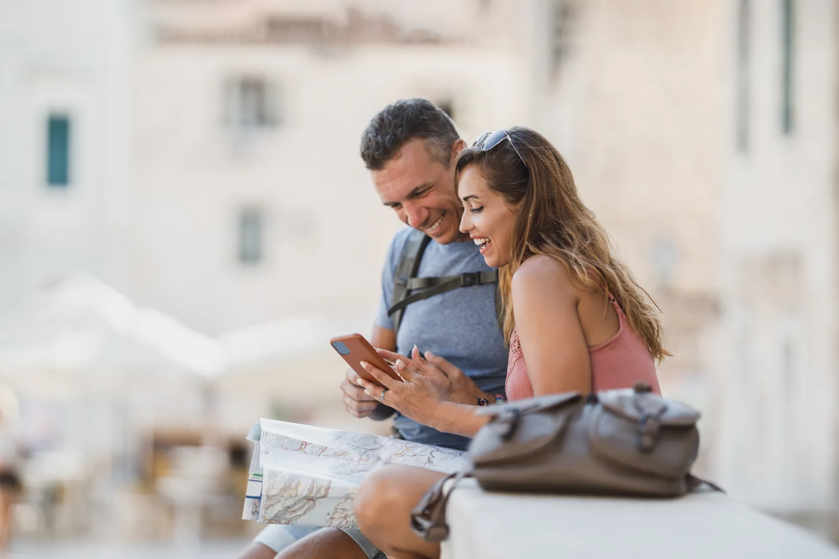 Ein lächelndes Paar sitzt auf einer Mauer und betrachtet gemeinsam ein Smartphone. Eine Stadtkarte liegt neben ihnen.