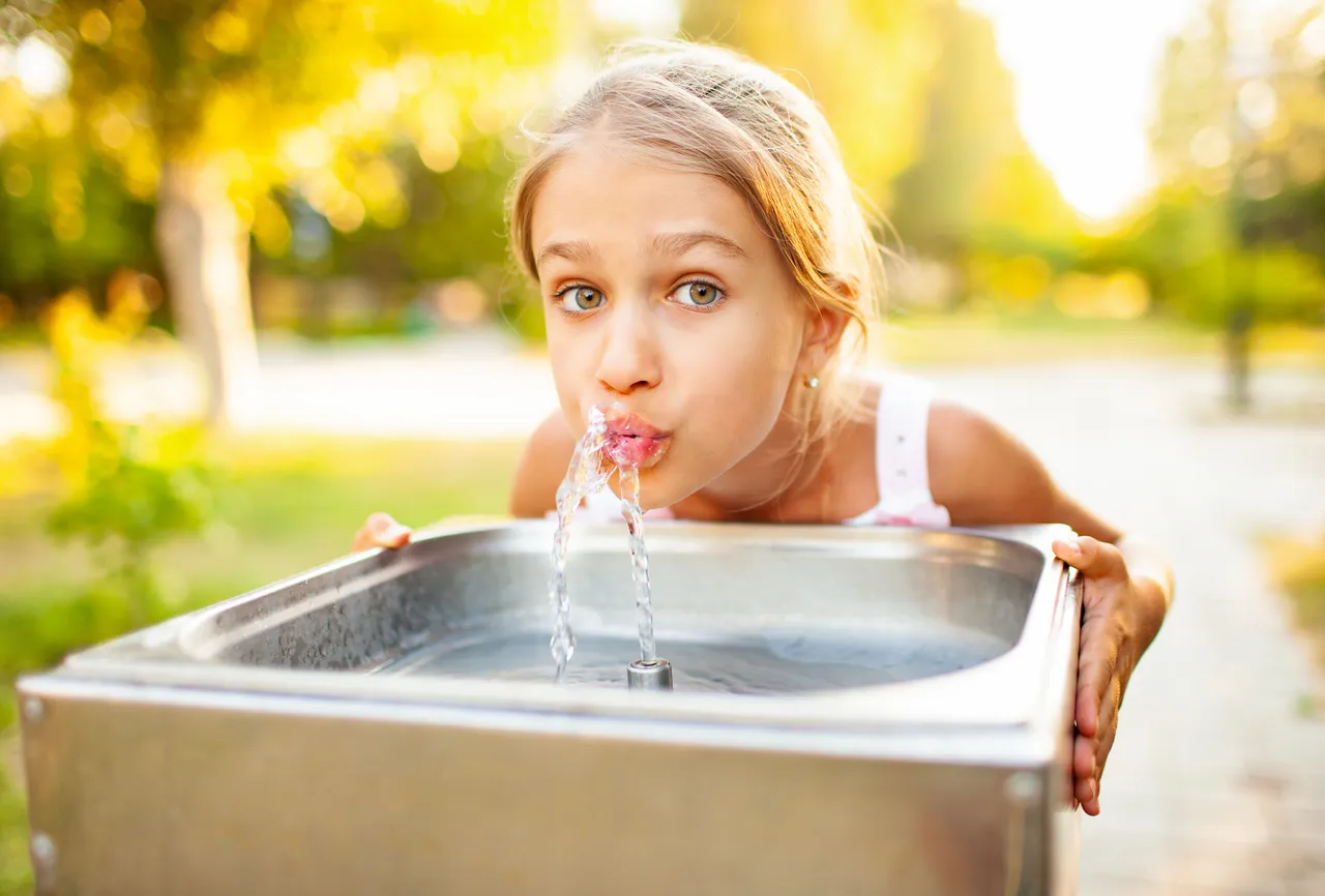 Ein blondes Mädchen trinkt aus einem öffentlichen Trinkwasserbrunnen im Freien. Der Hintergrund ist unscharf und zeigt Bäume und Grünflächen.