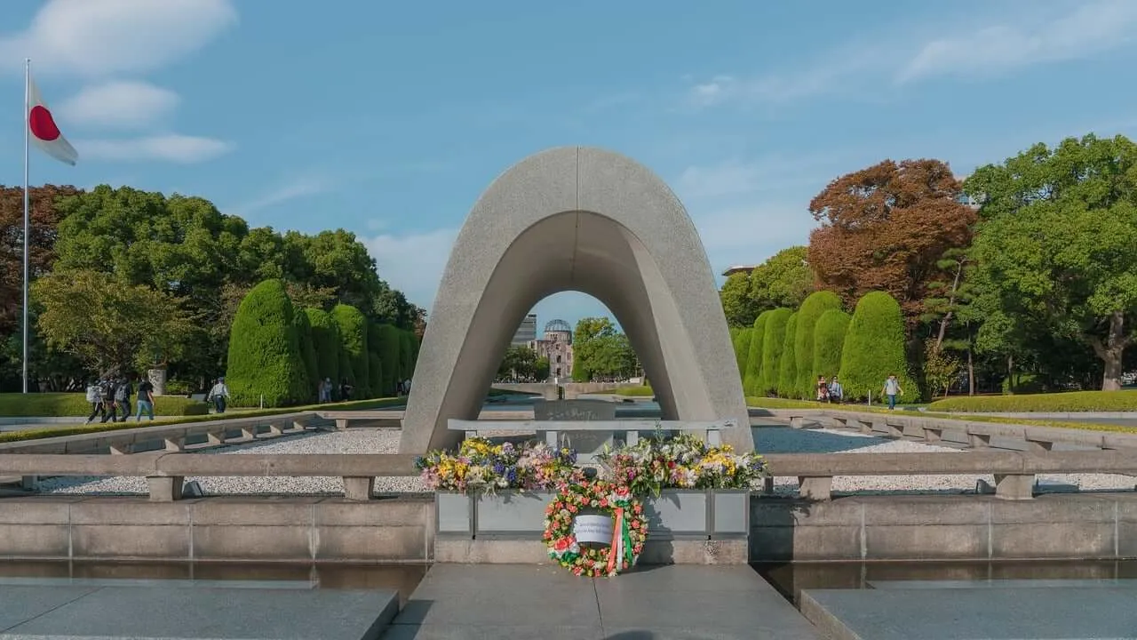 Gedenkstätte im Hiroshima Peace Park mit einem steinernen Bogen, durch den das Atombomben-Dome-Museum zu sehen ist. Vor der Anlage Blumenkränze.