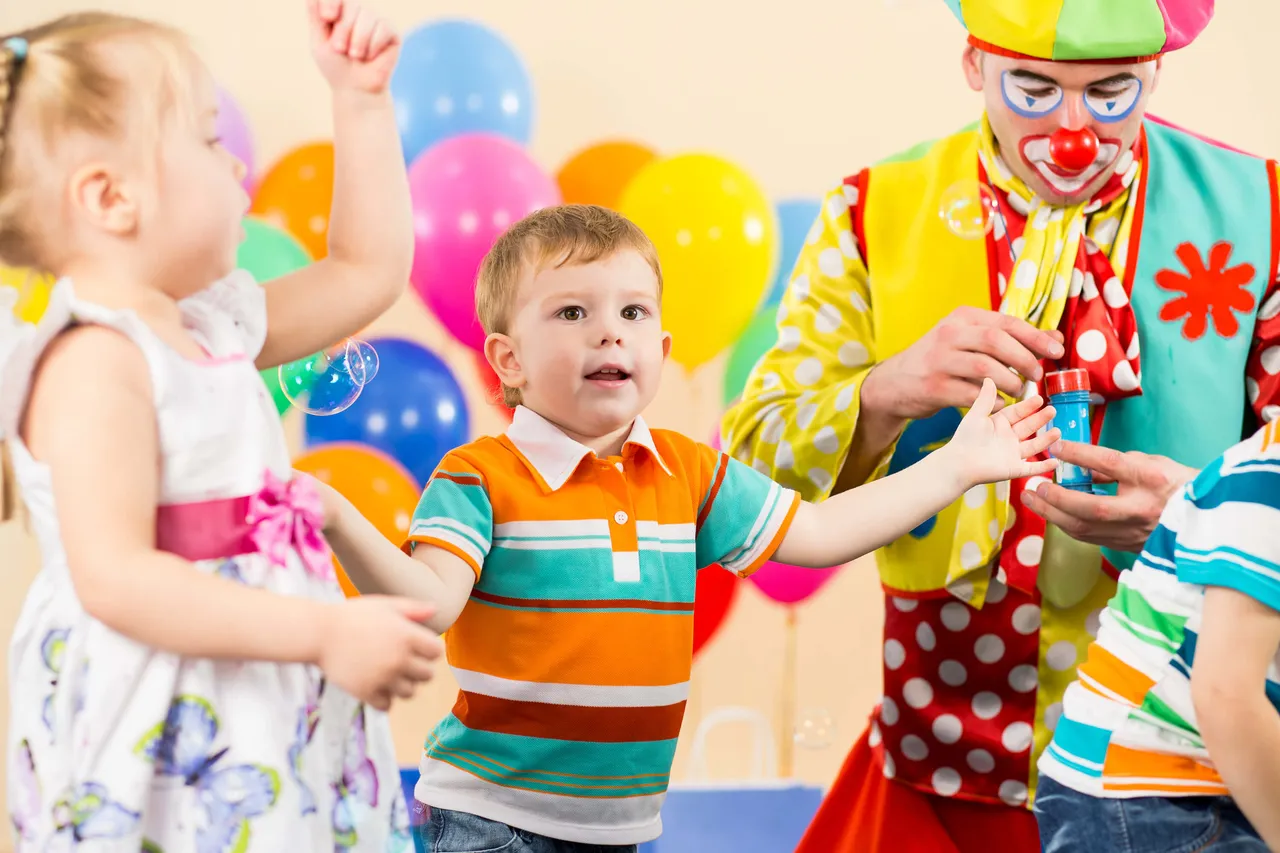 Ein Clown macht Seifenblasen für zwei Kinder auf einer Geburtstagsfeier. Im Hintergrund sind bunte Luftballons zu sehen.