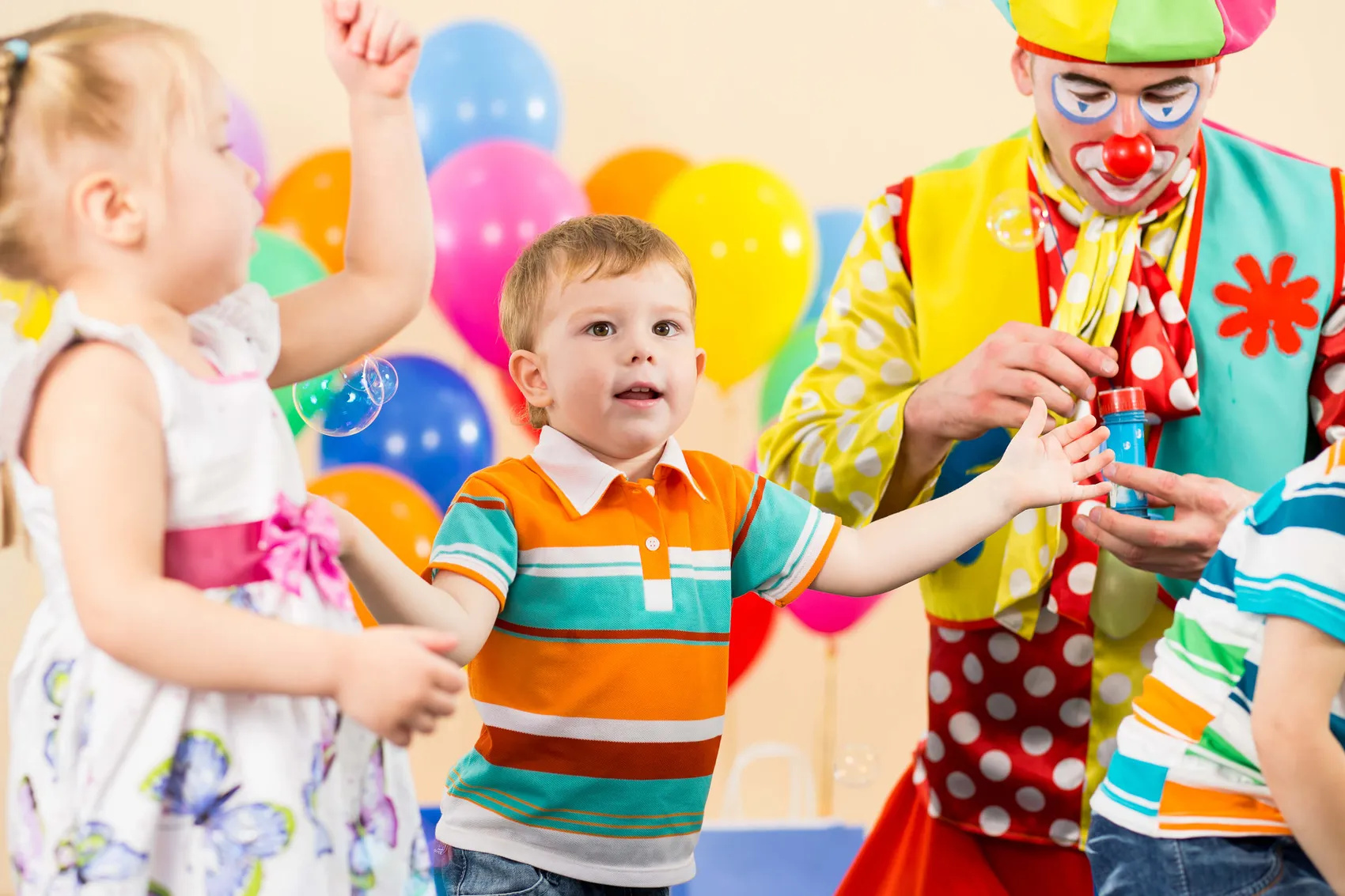 Ein Clown macht Seifenblasen für zwei Kinder auf einer Geburtstagsfeier. Im Hintergrund sind bunte Luftballons zu sehen.