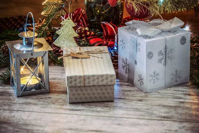 Ein brennendes Windlicht und zwei verpackte Geschenke unter den Zweigen eines geschmückten Weihnachtsbaums auf einer Holzoberfläche.