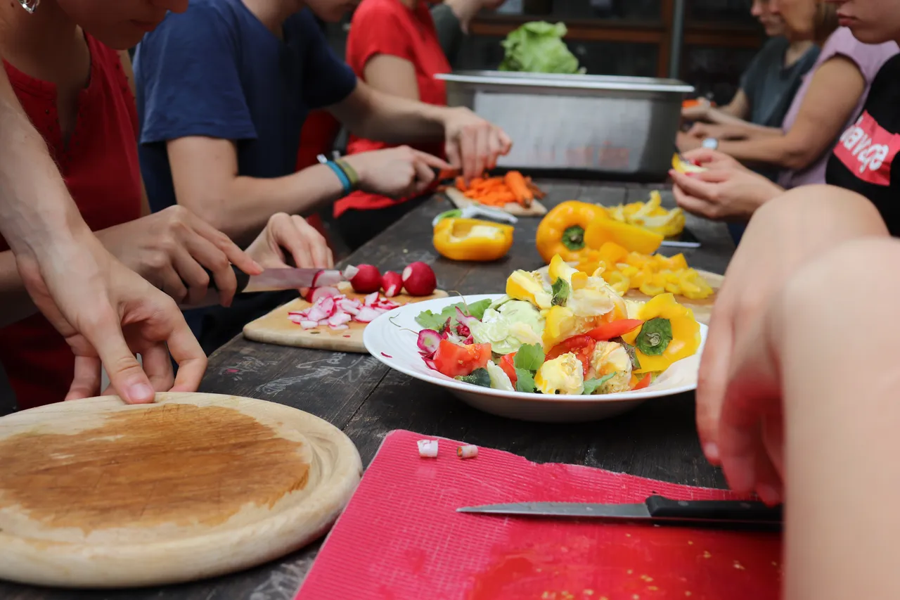 Mehrere Personen bereiten gemeinsam verschiedenes Gemüse wie Paprika und Radieschen auf einem Holztisch vor.