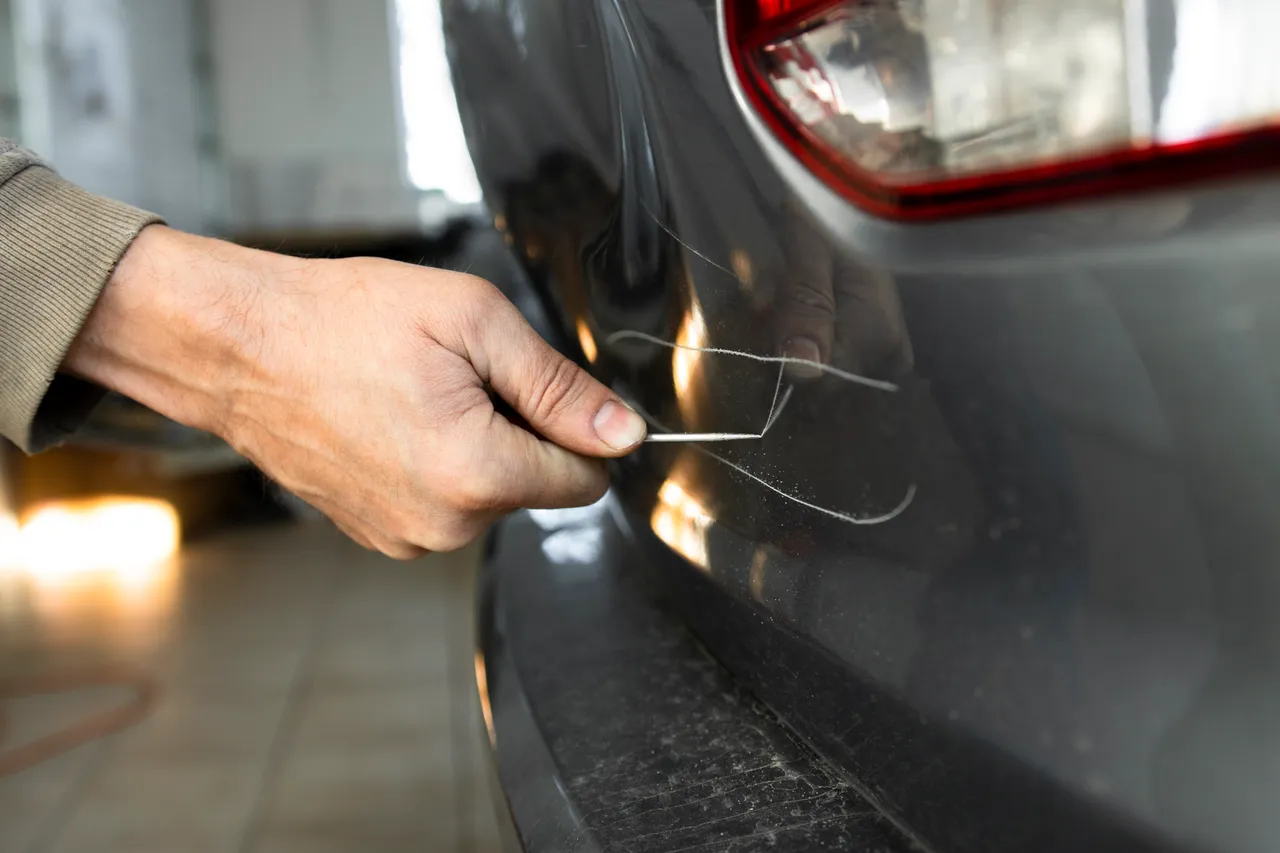 Eine Hand entfernt mit einem Metallwerkzeug eine Beschädigung an der Lackierung eines grauen Autos. Das Bild illustriert Vandalismus.