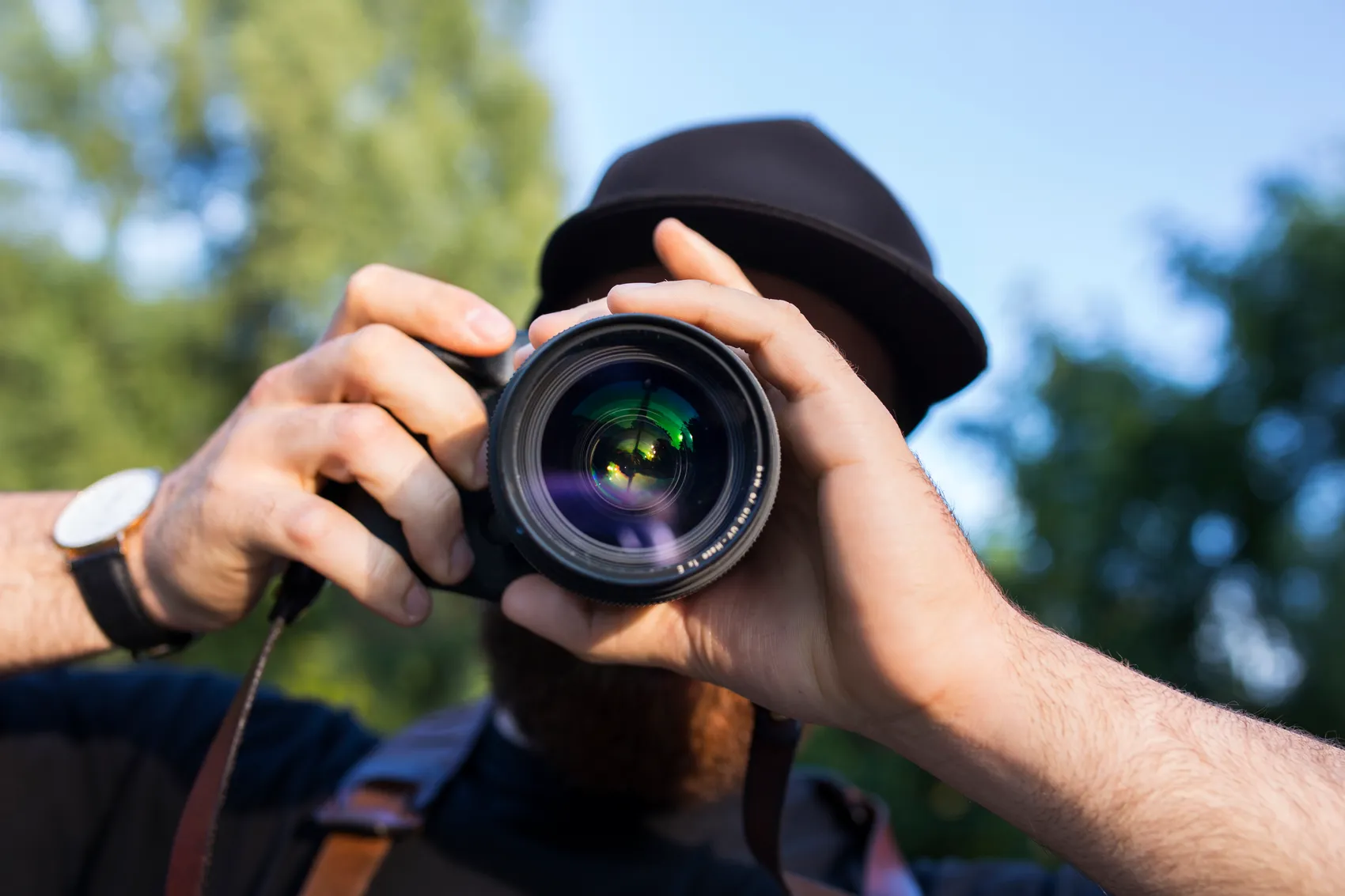 Nahaufnahme eines Mannes mit schwarzem Hut, der eine Kamera mit großem Objektiv direkt in die Linse hält.
