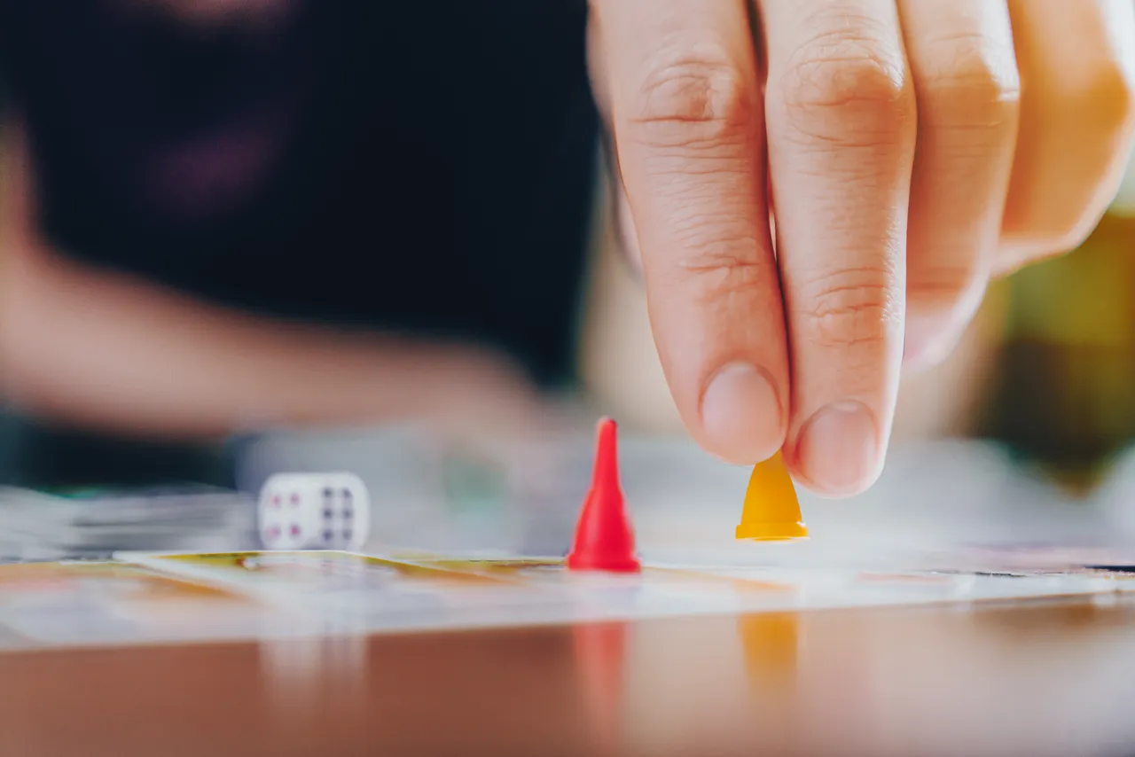 Eine Hand bewegt eine gelbe Spielfigur auf einem Brettspiel neben einer roten Spielfigur und einem Würfel.