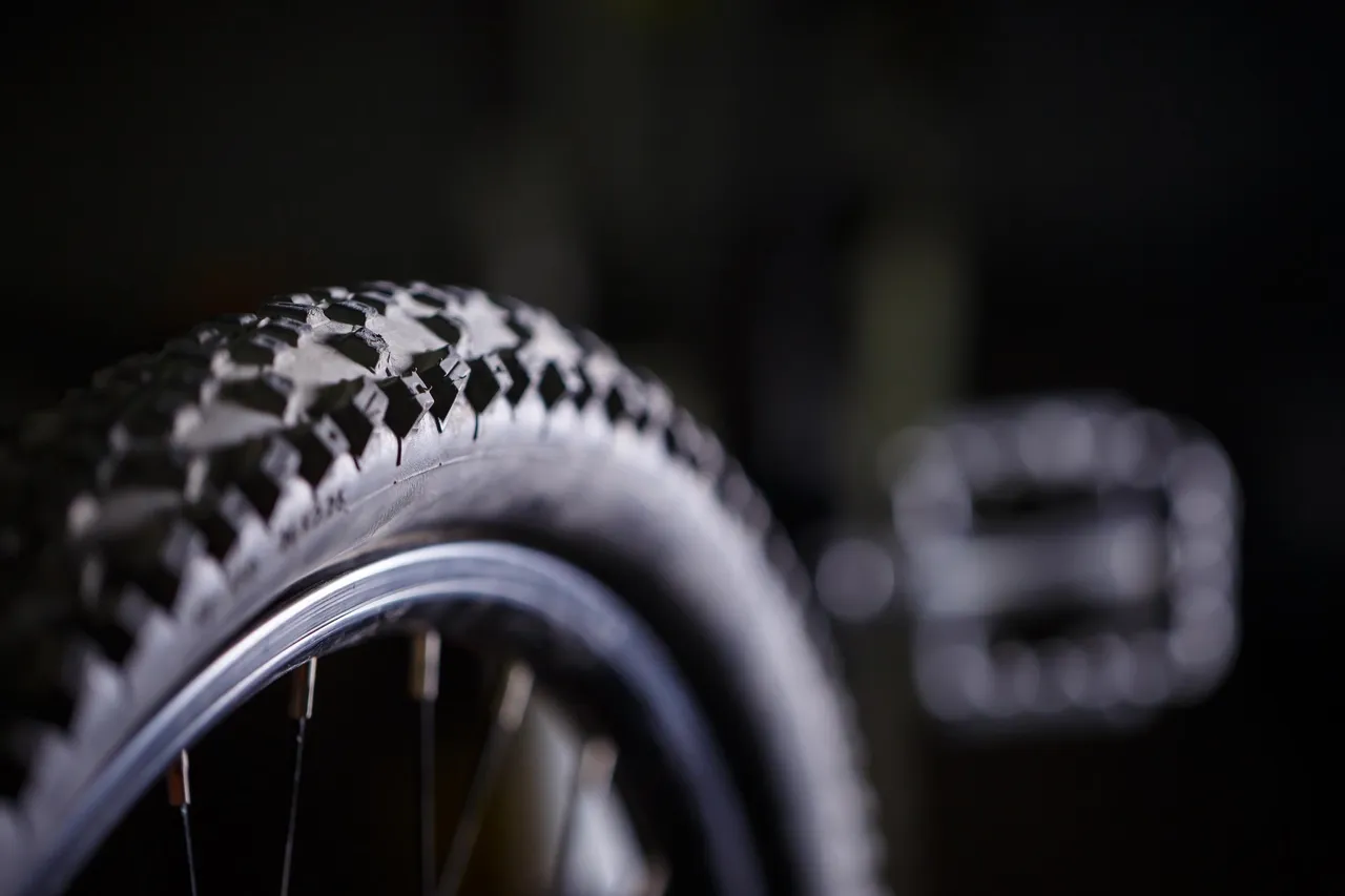 Nahaufnahme eines Mountainbike-Reifens mit grobem Profil und einem Pedal vor dunklem Hintergrund.